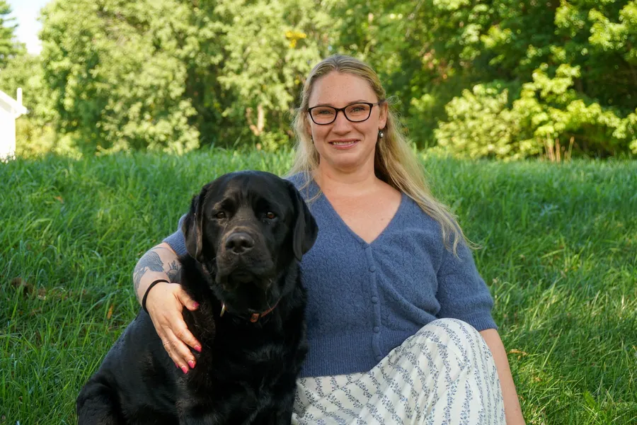 Marjorie Hunt, NP, sitting outside on the grass with her black Labrador